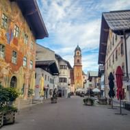 Voetgangersstraat in Mittenwald met kleurrijk beschilderde gebouwen en barokke kerktoren. Plantenbakken en terrassen langs de straat onder blauwe lucht.