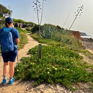 Wandelaar met blauwe rugzak op zandig kustpad in Portugal, omringd door groene vegetatie en agaves, met zeekliffen zichtbaar.