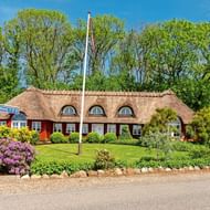 Traditioneel Deens rieten huis met rood-witte gevel, boogvensters en kleurrijke tuin met roze bloemen. Deense vlag op paal ervoor.