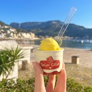 Hand houdt sinaasappelijs-bekertje met lepel vast tegen achtergrond van Port de Sóller strand met baai, bergen en palmbomen.