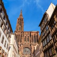 Gotische kathedraal van Straatsburg met sierlijke spits gezien vanaf smalle straat met traditionele vakwerkhuizen onder blauwe lucht.