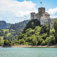 Burcht Czorsztyn op een beboste heuvel boven het turquoise Czorsztyn-stuwmeer, met groene bergen op de achtergrond onder een blauwe lucht.