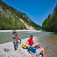Twee fietsers rusten op een kiezelbank naast de heldere turquoise Isar, omringd door beboste bergen onder blauwe lucht.