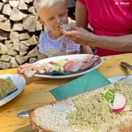 Familie snack in Koppental met Steirerkas spread op donker brood, gegarneerd met radijsschijfjes. Vleeswaren en gebak op houten tafel.