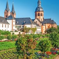 Kloostercomplex Seligenstadt met barokke kerktorens en koepel tegen blauwe lucht. Kleurrijke bloemperken en groene tuinen op voorgrond.
