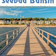 Houten pier die de Oostzee in loopt bij Bansin, Usedom. Kleurrijke gebouwen langs het zandstrand onder blauwe lucht met herfstbomen.