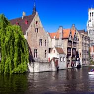 Middeleeuwse gebouwen langs een gracht in Brugge met het Belfort. Een treurwilg hangt over het water, een bootje met toeristen vaart voorbij.