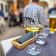 Glas witte wijn op een restauranttafel met wazige gasten op de achtergrond. Een houten dienblad met servet en waterglas zijn dichtbij.