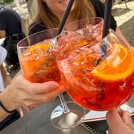 Drie handen houden grote wijnglazen vast gevuld met oranje Aperol Spritz-cocktails, ijsblokjes en sinaasappelschijfjes, proostend op een terras.