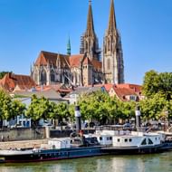 De gotische Dom van Regensburg met tweelingtorens domineert de skyline boven rood-bedekte gebouwen. Een passagiersboot vaart op de Donau.