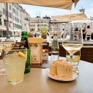 Buitentafel met witte wijnglazen, cocktail, groene bierfles en brood op Piazza Giacomo Matteotti in Udine met historische gebouwen.