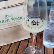 Wijnglas gevuld met troebele Federweisser op houten tafel met papieren zak 'Zum grünen Kranz' en waterglas op de achtergrond.
