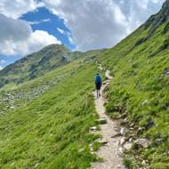Wandelaar met blauwe rugzak op smal bergpad in Nationaal Park Hohe Tauern, omringd door groene hellingen onder bewolkte hemel.