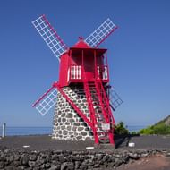 Traditionele rode molen met witte wieken op vulkanische rotskust op de Azoren. Stenen basis met houten structuur tegen blauwe lucht en oceaan.
