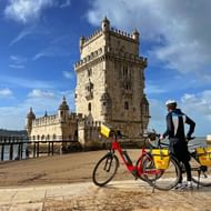 Fietser met rode toerfiets staat naast het fort Torre de Belém in Lissabon onder blauwe hemel met witte wolken.