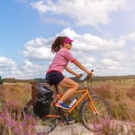 Vrouw fietst door paars bloeiende heidevelden in Nationaal Park De Veluwe met toerfiets en fietstassen onder bewolkte hemel.