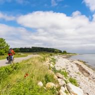 Fietser in oranje jas op kustpad bij Bøgebjerg Strand, Denemarken. Het pad loopt tussen groene weiden en een rotsachtig strand met kalme zee.