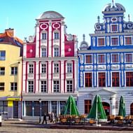 Rij kleurrijke barokgebouwen op het oude stadsplein van Szczecin met rode, blauwe en gele gevels. Groene parasols beschaduwen terras.