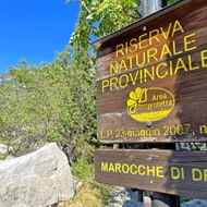 Bruin houten bord voor provinciaal natuurreservaat Marocche di Dro met gele tekst, omgeven door groene vegetatie en rotsen onder blauwe lucht.