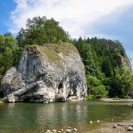 Grote witte kalksteenformatie met grotten rijzend uit de Dunajec, bedekt met groene bomen onder een blauwe lucht met wolken.