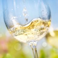 Close-up van witte wijn die dynamisch spat in een wijnglas tegen een wazige buitenachtergrond met groene vegetatie en blauwe lucht.