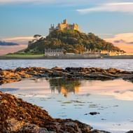 St Michael's Mount getijdeneilend met middeleeuws kasteel en kerk op heuveltop, omringd door water bij zonsondergang. Rotsachtige kustlijn.