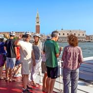 Passagiers op het zonnedek van MS Ave Maria kijken naar de skyline van Venetië met de Campanile en het Dogenpaleis over de lagune bij blauwe lucht.