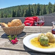 Drie Zuid-Tiroolse knödels op wit bord met kruidensaus, broodmand, wijnglas en kan op houten tafel met bergzicht.