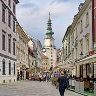 Voetgangersstraat in het historische centrum van Bratislava met kleurrijke barokgebouwen, terrasjes en een barokke klokkentoren op de achtergrond.