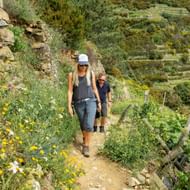 Twee wandelaars op een smal bergpad in Cinque Terre, omringd door wilde bloemen en terrasvormige wijngaarden op steile hellingen.