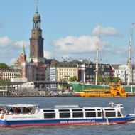 Witte passagiersboot op de Elbe in Hamburg met historische St. Michaelis kerktoren en kleurrijke havengebouwen op achtergrond.