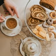 Handen houden koffiekop naast glazen bord met Sloveens gebak waaronder potica rolcake, walnootkoekjes en diverse traditionele zoetigheden.