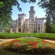 Wit neogotisch kasteel met cilindrische toren gezien door boomtakken, met formele tuinen en rond bloembed op de voorgrond.
