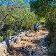 Vrouwelijke wandelaar met blauwe rugzak op rotsachtig gravelpad door mediterraan bos met dichte groene vegetatie en blauwe lucht.
