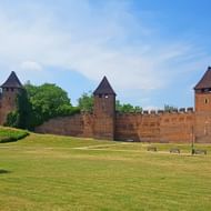 Middeleeuwse bakstenen vestingmuren met ronde torens en kantelen in Nymburk, omgeven door groen gazon en bomen onder blauwe lucht.