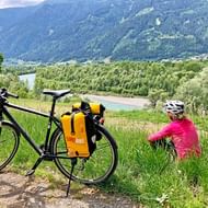 Fietser in roze jas zit in gras naast toerfiets met gele fietstassen, uitkijkend over Drau-vallei met beboste bergen.