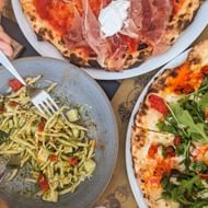 Twee pizza's met prosciutto, mozzarella en rucola, en een pastagerecht met pesto op een houten tafel in Piëmont-Ligurië.