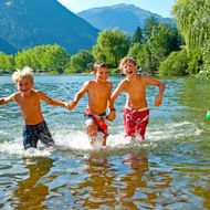 Drie jongens hand in hand rennend Lake Zell Maishofen in, spetterend water. Een meisje zit op groene opblaasbare. Bergen en bomen op de achtergrond.