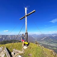 Vrouwelijke wandelaar staat naast een houten topkruis op de Kalmberg-piek met panoramisch uitzicht op bergen en dal.
