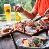 Persoon snijdt traditionele Beierse worst en vleeswaren op houten plank, met bierglas en krakeling op buitentafel in Tegernsee-regio.