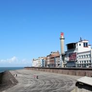 Kustpromenade in Vlissingen met witte vuurtoren, kleurrijke gebouwen langs de waterkant en mensen wandelend op brede stenen pier onder blauwe lucht.