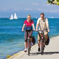 Man en vrouw fietsen op pad langs het Bodenmeer met blauw water, zeilboten en bergen op de achtergrond onder heldere hemel.