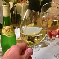 Hand houdt glas witte wijn vast tijdens proeverij, met groene wijnfles en andere glazen op tafel in restaurant setting.