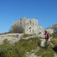 Twee mountainbikers op een onverharde weg naderen een oude stenen vesting in Montenegro, met rotsachtige bergen en blauwe lucht op de achtergrond.