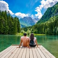 Twee personen zitten op houten steiger met uitzicht op turquoise meer in Slovenië, omringd door beboste bergen en dramatische lucht.