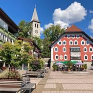 Saalfelden centrum met kleurrijke gebouwen, kerktoren met klok, buitenterras en plantenbakken op geplaveide plein onder blauwe lucht.