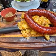 Krainer worst in hoefijzervorm op een houten plank met gebakken aardappelen, vergezeld van een klein schaaltje kruiden en een mes.