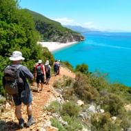 Groep wandelaars met rugzakken op een rotsachtig pad langs de Albanese kust, uitkijkend over turquoise zee en strand.
