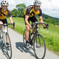 Twee wielrenners in geel-zwarte shirts op racefietsen op asfaltweg door groen landschap met heuvels en bossen.