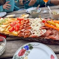 Houten plank met Oostenrijkse worst, kaas, groenten en brood op rustieke tafel. Mensen delen maaltijd met bloemenservies.